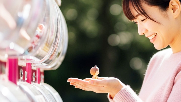 30代日本人女性がガチャガチャを楽しんでいる様子。欲しかった品をゲットして嬉しそう。大人の趣味としての共感と秘密の楽しみを表現。